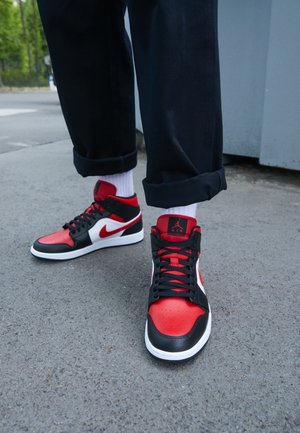 Person iført sorte, røde og hvide Nike Air Jordan high-top sneakers med hvide sokker og sorte bukser, stående på asfalt.