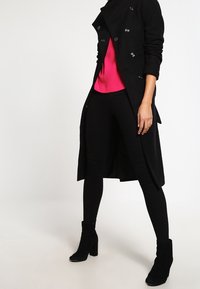 Cappotto nero doppiopetto con bottoni argento, indossato sopra un top rosa fluo e leggings neri, abbinato a stivaletti neri alla caviglia.