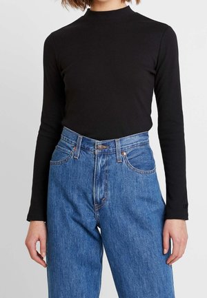 Henkilö pukeutuneena istuvaan mustaan pitkähihaiseen yläosaan, joka on tungettu korkeavyötäröisiin sinisiin farkkuihin, joissa on etutaskut ja vyölenkit.