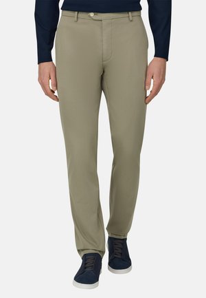 Man die beige slim-fit broek met lussen voor een riem en marineblauwe sneakers draagt, staand tegen een effen achtergrond.