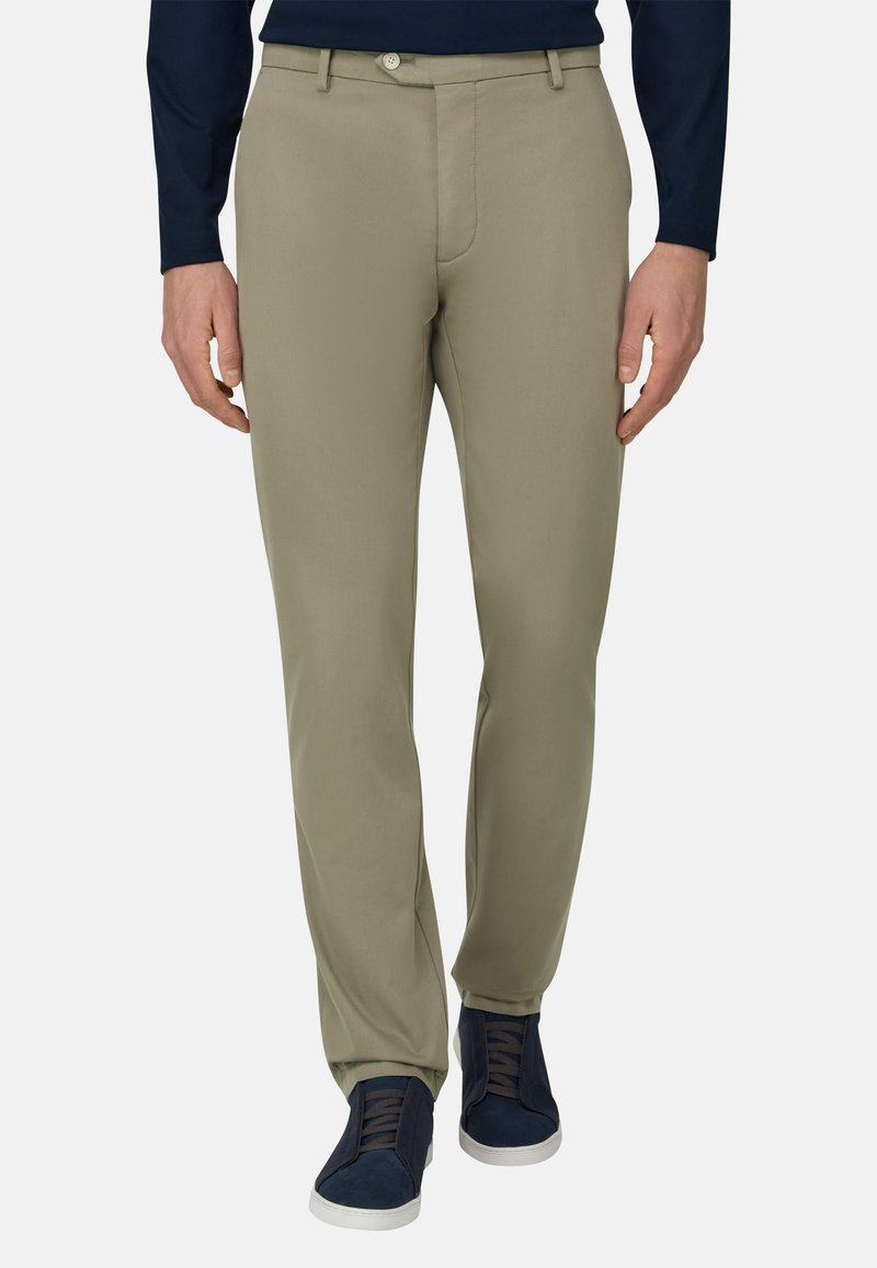 Homme portant un pantalon beige slim avec passants de ceinture et des baskets bleu marine, debout devant un fond uni.