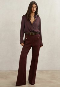 Burgundy silk shirt with buttoned collar, paired with high-waisted burgundy trousers featuring large pockets and gold button accents.