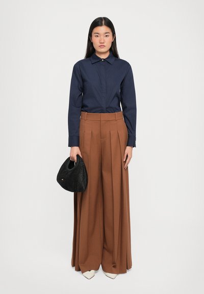 Navy collared shirt, brown wide-leg pleated trousers, black woven handbag, and white pointed-toe shoes on a plain background.