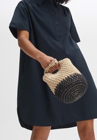 Personne portant une robe noire à manches courtes, tenant par la poignée un petit sac rond tissé avec des blocs de couleur beige et noir.