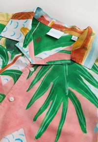 Camisa de botones de manga corta en tejido ligero con estampado de hojas de palma verdes sobre un fondo rosa, con acentos de contraste en azul y marrón.