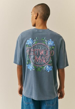 Person med kort hår som vänder sig bort, bär en blekt blå Fleetwood Mac t-shirt med blommönster och blå jeans.