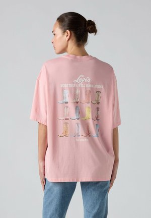 Nainen nutturalla, pukeutunut vaaleanpunaiseen Levi's t-paitaan, jossa on erilaisia värikkäitä cowboy-saappaita ja tekstiä selässä, yhdistetty sinisiin farkkuihin.