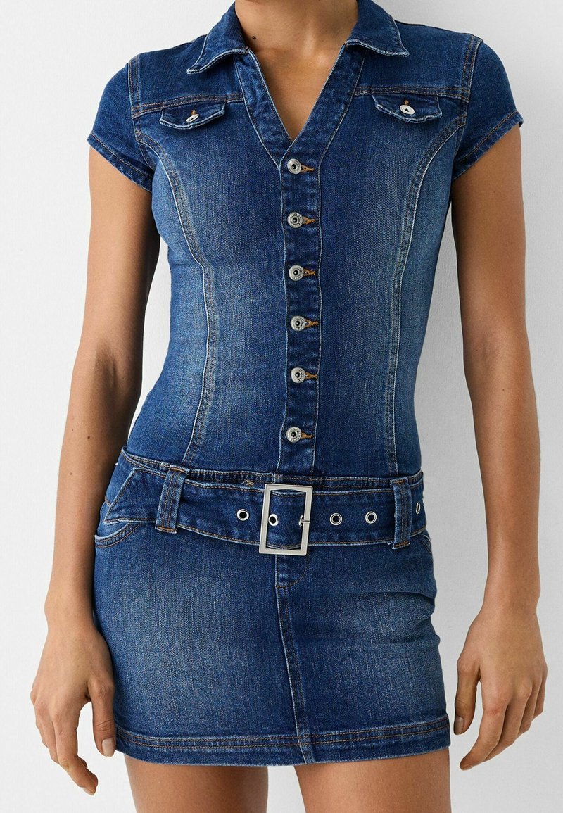 Žena v přiléhavých džínových šatech s krátkým rukávem, knoflíkovým zapínáním vpředu, límcem, kapsami na prsou a odpovídajícím páskem se čtvercovou přezkou.