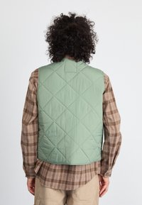 Zaļš, quiltēts veste ar augstu apkakli un rombu rakstu, uzvilkta virs brūnas sarkanās svītras garām piedurknēm krekla. Gluda tekstūra, ikdienas dizains.