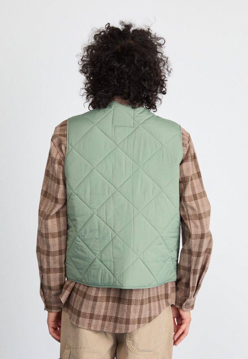 Quilted grøn vest med høj krave og diamantmønster, båret over en brun ternet skjorte med lange ærmer. Glat tekstur, afslappet design.