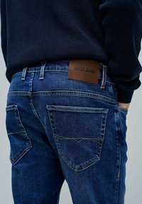 Plave traperice s izblijedjelim izgledom, s stražnjim džepovima, žutim šavovima i smešnim kožnim etiketom s natpisom "SALSA JEANS."