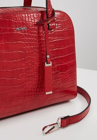 Sac à main rouge en cuir synthétique effet crocodile, de forme arrondie, équipé de ferrures argentées, d'une bandoulière réglable et d'une étiquette portant le nom de la marque.