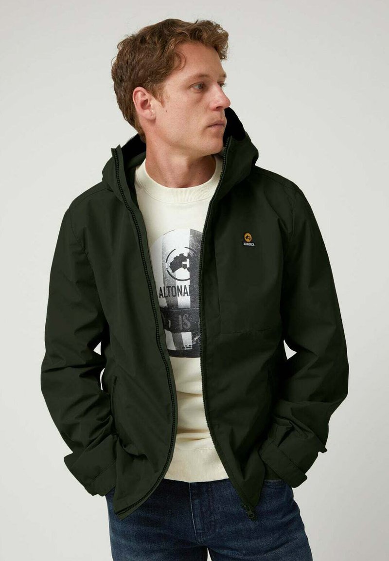 Chaqueta de cremallera verde oscuro con capucha, que presenta un pequeño logo en el pecho y un bolsillo frontal. Se lleva sobre una sudadera gráfica de color claro.