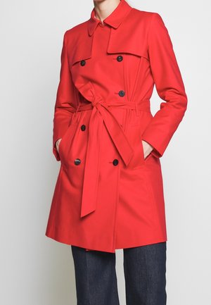 Rød dobbeltradet trenchcoat med sorte knapper og bindebælte i samme stof, båret over mørkeblå jeans, hænder i frakkens lommer.