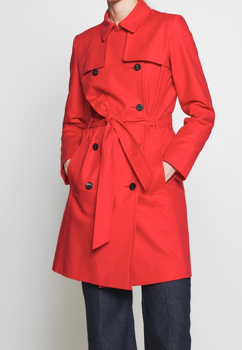 Trench-coat rouge croisé avec boutons noirs et ceinture à nouer, porté sur un jean bleu foncé, mains dans les poches du manteau.