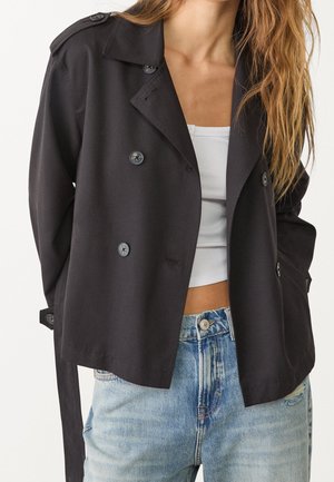 Woman wearing a black double-breasted jacket, white crop top, and blue high-waisted jeans with buttons visible.