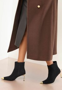 Fekete velúr bokacsizmák arany hegyes orr díszítéssel, magas stiletto sarkakkal és oldalsó cipzárral, barna kabáttal párosítva.