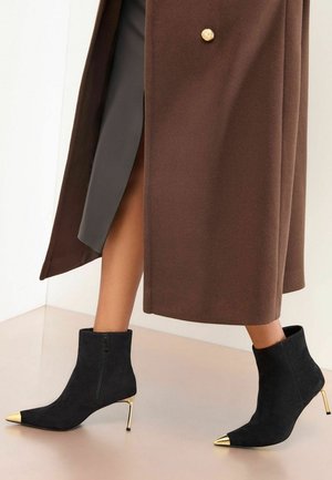 Melni zamšaini potītes zābaki ar zelta norādītām purngalu akcentam, augstām stiletto papēžiem un sānu ķēdēm, kombinēti ar brūnu mēteļu.