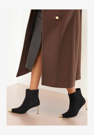 Schwarze Wildleder-Stiefeletten mit goldenen, spitzen Zehenspitzen, hohen Stilettos und seitlichen Reißverschlüssen, kombiniert mit einem braunen Mantel.