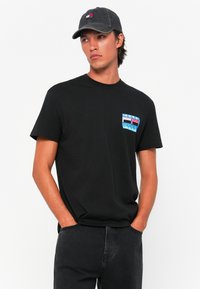 Schwarzes Baumwoll-T-Shirt mit einem kleinen, mehrfarbigen Logo-Patch auf der linken Brust. Kombiniert mit dunkelgrauen Jeans und einer passenden Kappe.