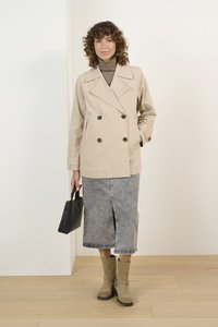 Chaqueta beige de doble botonadura, falda de mezclilla gris con abertura frontal, bolso negro y botas de tobillo beige con detalles decorativos.