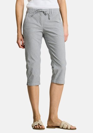 Frau trägt hellgraue, gekürzte Hose mit Bindeband in der Taille und weiße Slippersandalen, steht vor schlichtem Hintergrund.