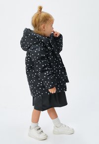 Puffjakke i sort med hvide prikker, med hætte, quiltet design og knaplukning foran, båret over en sort nederdel og hvide sneakers.