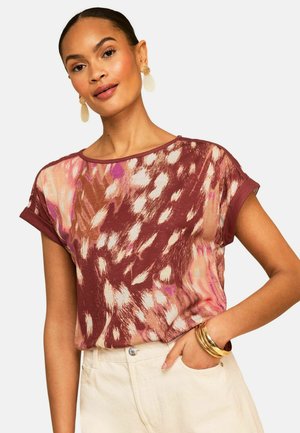 Naine, kes kannab lühikeste varrukatega, kahvatu punase ja beežika abstraktse mustriga pluusi keeratud varrukate ja kreemjate pükstega, kuldsete rõngakujuliste kõrvarõngaste ja kuhjatud käevõrudega.