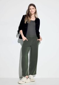 Zwarte textuur cardigan, grijzige tanktop, olijfgroene broek met een elastische band waarop "POSITIVITY" is gedrukt, en witte sneakers met zwarte accenten.