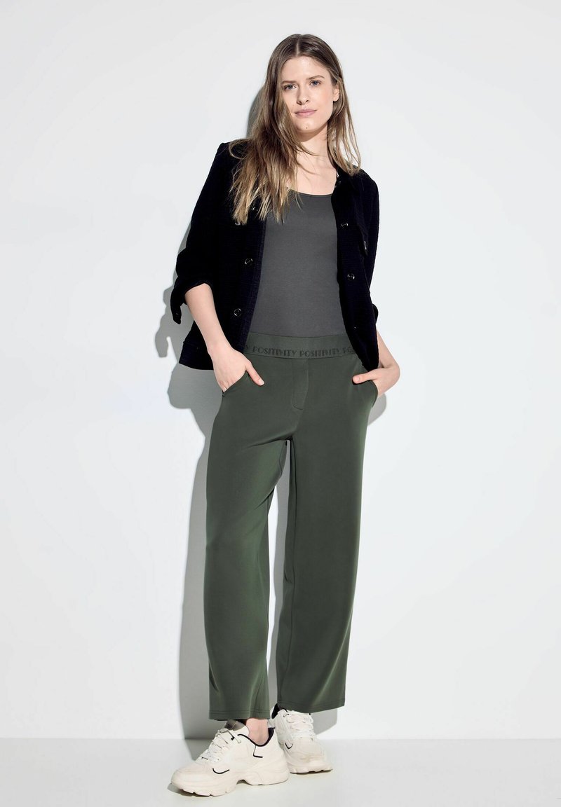 Zwarte textuur cardigan, grijzige tanktop, olijfgroene broek met een elastische band waarop "POSITIVITY" is gedrukt, en witte sneakers met zwarte accenten.