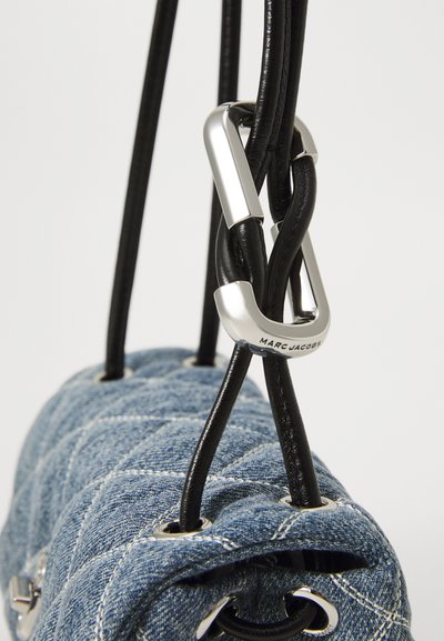 Prim-plan al unei genți din denim bleumarin, matlasate, cu șnururi din piele neagră trecute prin ochiuri argintii și un închizător argintie gravată "Marc Jacobs."
