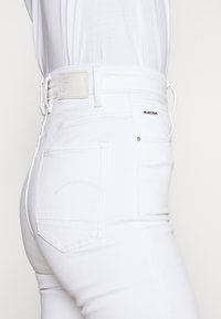 Jeans de mezclilla blanca con un diseño ajustado. Presenta un parche de metal con el logo en la cintura y detalles de costura en el bolsillo trasero. Textura suave.