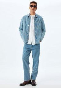 Giacca in denim azzurro chiaro con tasca, camicia bianca a bottone, jeans azzurri chiari e stivali marroni scuri, completati da occhiali da sole neri.