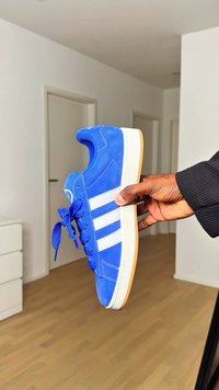 Kék velúr sportcipő fehér csíkkal, kerek orral és kék fűzővel. Gumitalppal rendelkezik, amelyen bézs díszítés található. Kézben tartva.