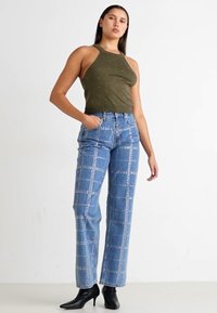 Top halter verde oliva, abbinato a jeans in denim blu con motivo a griglia borchiato e taglio dritto. Stivali neri con punta a punta completano il look.