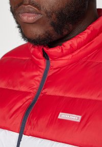 Rote Daunenjacke mit hohem Kragen, versehen mit grauen Akzenten und einem sichtbaren Etikett "JACK & JONES." Glatte Textur und gestepptes Design.