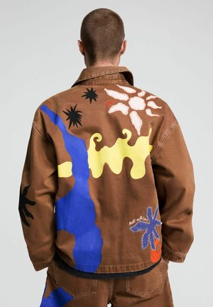 Mann mit brauner Jacke mit abstrakten bunten Formen in Blau, Gelb, Schwarz, Weiß und Orange steht mit dem Rücken zu einem einfarbigen Hintergrund.