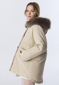 Beige gewatteerde jas met een doorgestikt patroon, voorzien van een nepbontcapuchon, een taille die is getailleerd en zijzakken. Gladde textuur, knielange ontwerp.