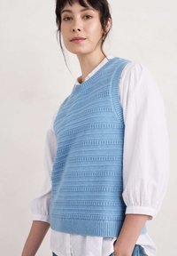 Gilet en tricot bleu clair avec des rayures horizontales texturées, encolure ronde et ourlet côtelé, porté par-dessus une chemise à manches longues blanche.