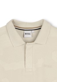 Polo shirt beige chiaro con un motivo camo testurizzato, caratterizzato da un colletto a costine e un'apertura con due bottoni. Logo ben visibile.