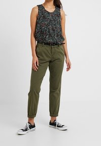 Top senza maniche con stampa floreale e dettagli arricciati in nero, rosso e verde, abbinato a pantaloni a sigaretta verde oliva e sneakers in tela nere.