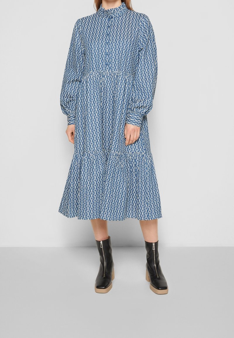 Femme portant une robe bleue mi-genou avec un motif floral blanc, manches longues et col montant, associée à des bottines noires à fermeture éclair.
