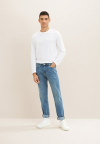 Bela dolga majica z dolgimi rokavi, modri jeans s podravljenimi manšetami in bele teniske. Model stoji seno na eni roki v žepu ob svetlem ozadju.
