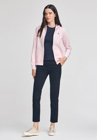 Felpa con zip di un delicato rosa chiaro, con tasche frontali, abbinata a una maglietta navy, pantaloni scuri e sneakers beige; presenta un cappuccio con stringa e dettagli del logo.