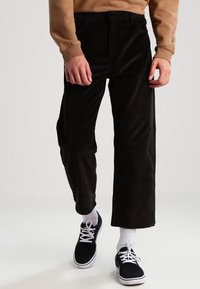 Svarta kordbyxor med vida ben och texturerad tyg. Bärs tillsammans med en brun sweatshirt och svarta sneakers med vita snören och strumpor.