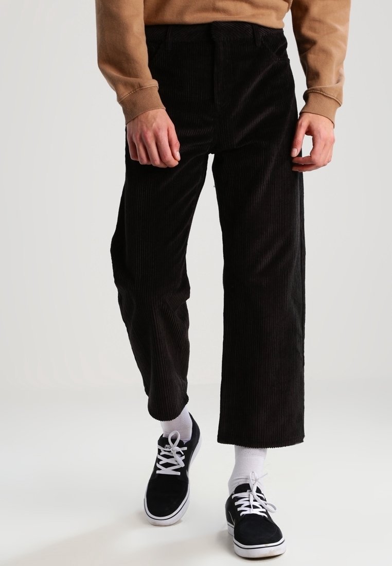 Svarta kordbyxor med vida ben och texturerad tyg. Bärs tillsammans med en brun sweatshirt och svarta sneakers med vita snören och strumpor.