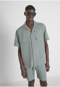 Lichtgroen, gestructureerd shirt met korte mouwen en twee borstzakken, gecombineerd met bijpassende shorts. Het model draagt grote, hoekige zwarte zonnebrillen.