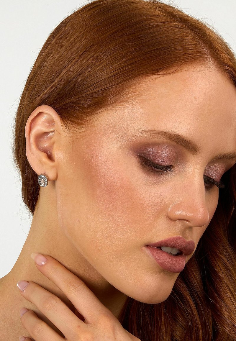 Zilveren studs met een geometrisch ontwerp, geplaatst tegen een lichte huidtoon en lang, golvend roodan. Zachte, neutrale make-up benadrukt de kenmerken.