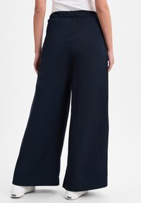 Marineblauwe wijde broek met een elastische tailleband, vervaardigd uit gladde stof en met een losse pasvorm. De schoenen die onderaan zichtbaar zijn, zijn wit met zwarte accenten.