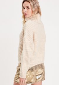 Pull en tricot beige avec un col haut, un motif texturé et une coupe oversize, associé à un short en sequins dorés brillants.